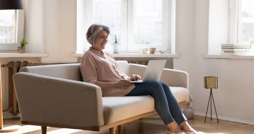 Image of smiling senior woman sitting on couch using laptop at home to attend Virtual IOP